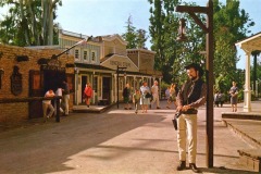 A man dressed as a cowboy leans against a wooden post in Frontier Village, a Western-themed town in San Jose, California, with people strolling by the general store and saloon under sunny skies.