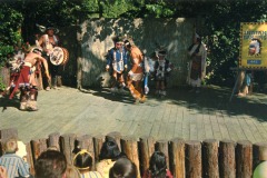 A group of people in traditional Native American clothing perform a dance on an outdoor wooden stage at Frontier Village in San Jose, California, watched by an audience. A sign reading Indian Dancing is displayed on the right.