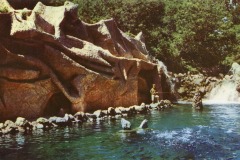 A rocky grotto with cave-like openings borders a clear blue pool, surrounded by lush green trees. Reminiscent of California’s old Frontier Village near San Jose, a small waterfall flows at the far end and a person stands near the cave entrance.