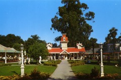 A vintage photo of Frontier Village theme park entrance in San Jose, California, featuring a red-roofed clock tower, green lawns, flowers, people walking, and lamp posts under a clear blue sky.