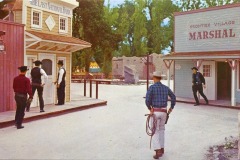 Four men dressed as cowboys stand on a wooden street in Frontier Village, an Old West town in San Jose, California. One heads toward a bank, another toward a marshal’s office, and two stand near a building. A tipi and trees are visible in the background.