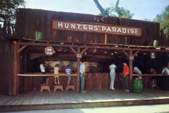 A vintage shooting gallery called Hunters Paradise at Frontier Village in San Jose, California, with people aiming at targets. Children and adults stand at wooden counters under a rustic sign, surrounded by trees and decorative props.