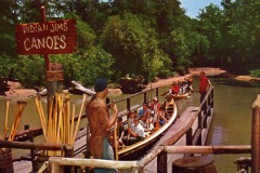 A group of people sit in long canoes on a river at Frontier Village in San Jose, California. Guided by leaders, they glide past a rustic wooden dock under a sign reading Indian Jims Canoes, surrounded by lush trees and greenery.
