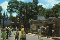 People walk past a rustic “Mexican Market” building at Frontier Village in San Jose, California, with vendors and colorful goods outside under a bright blue sky, surrounded by trees and other Western-style structures.