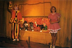 A clown in a colorful costume waves while standing next to a woman in a pink polka-dot dress at Frontier Village in San Jose, California. Behind them is a puppet stage with marionettes, set against a gold curtain.