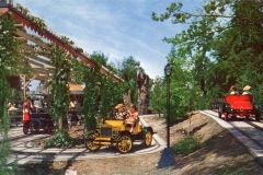 Children and adults ride vintage-style cars on winding tracks through a lush, green outdoor attraction at Frontier Village in San Jose, California. A pergola with people underneath is visible on the left beneath the sunny sky.