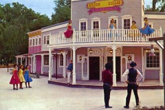 A vintage-style saloon with a wooden porch and balcony at Frontier Village in San Jose, California features women in colorful dresses posing, while three men dressed as cowboys stand and watch outside. The sign reads Silver Dollar Saloon.