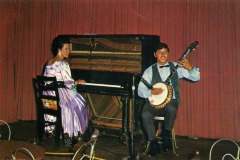 A woman in a purple dress plays a piano with its inner strings exposed, while a man in a vest and bow tie sits beside her playing a banjo. They perform onstage in front of red curtains at Frontier Village in San Jose, California.