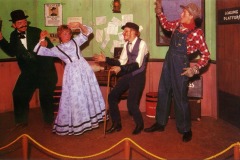 Four wax figures in frontier-era clothing reenact a dramatic scene at Frontier Village in San Jose: a woman in a blue dress appears startled as two men confront a plaid-clad man with a raised weapon near the “Loading Platform” sign.