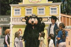 A person in a bear costume stands in front of a yellow Frontier Village building in San Jose, California, surrounded by six children and an adult, all outdoors on a sunny day.