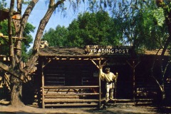 A rustic log cabin trading post is surrounded by trees at Frontier Village in San Jose, California. A statue of a person in Native American attire stands near the entrance, and a sign on the roof reads Trading Post.