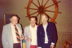Three men stand together, smiling in front of a large ships wheel mounted on the wall at Grotto Restaurant in Oakland, California. They are wearing suits and holding drinks, creating a casual and friendly atmosphere.