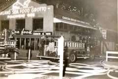 Vintage photo of a fire truck in front of the Oakland Sea Food Grotto restaurant near Jack London Square, California. Hoses are spread on the ground, and firefighters are visible. The buildings sign reads Charcoal Broiler Fish - Steaks - Chicken.