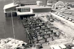 Black and white photo of a waterfront scene featuring a building labeled Grotto, a dock with dilapidated pilings, parked vintage cars, and a large ship in the background under a partly cloudy sky.