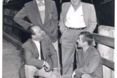 Four men in suits and casual shirts gather on a dock in Oakland, California, in front of a large ship. Two are standing, smiling, while two squat, looking up at the others as they enjoy a lively conversation reminiscent of an evening at the Grotto Restaurant.