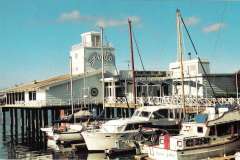 A marina with several boats docked in front of a building on stilts over the water. The building is white with a sign reading Guido, reminiscent of the charming settings around Jack London Square. A bright blue sky with a few clouds is visible in the background.