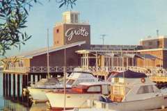 A wooden waterfront building labeled Grotto stands on stilts above the water. Several boats are docked nearby. The sky is clear, and sunlight illuminates the scene, with leafy branches visible in the foreground.