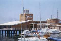 A waterfront building with the name Grotto on it stands on stilts. Several boats and yachts are docked in the foreground, and the sky is clear and blue.