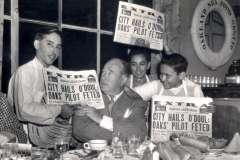 A group of men smiles and holds newspapers with the headline City Hails ODoul; Oaks Pilot Feted around a seated man in a suit at a table laden with food and drinks. In Californias own Oakland Sea Food Grotto, nestled by Jack London Square, a life preserver hangs on the wall behind them.