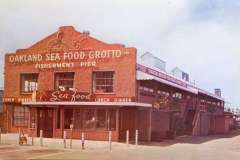 Historic building with Oakland Sea Food Grotto on the facade at Fishermens Pier. The structure is made of brick, featuring signage for seafood and lunch dinners, with an adjoining wooden staircase. A clear sky is seen in the background.