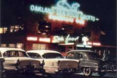 A vintage seafood restaurant in Oakland, California glows under neon signs reading Oakland Sea Food Grotto. Classic cars lined up front mirror the vibrant lights of this iconic Grotto Restaurant, capturing the charm of a bygone era.
