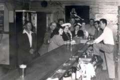A group of people gathered at a bar counter in Jack London Square, some seated and some standing, dressed in mid-20th-century attire. The bartender stands on the right, engaging with the patrons. A poster with a silhouette of a dancer is visible in the background, capturing Oaklands vibrant spirit.
