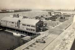 Black and white image of a waterfront scene at Jack London Square with a large industrial building labeled Canary Sea Foods Co. A few boats are docked nearby in Oakland, California, with an empty parking lot and a single car visible in the foreground.