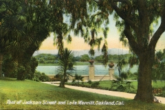 A scenic view of Lake Merritt, Oakland, California, from the foot of Jackson Street. The foreground features lush greenery with trees and plants. The lake is visible beyond a path, with mountains in the background under a colorful sky.