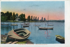 A serene view of boats anchored on Lake Merritt in Oakland, California, at dusk. Several boats are floating on the calm water, with trees and a small building visible along the shoreline under a pinkish-orange sky.
