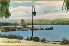 A vintage postcard image of Lake Merritt in Oakland, featuring a tranquil lake view with small boats docked along the shoreline. A string of lights is draped from a lamppost, and lush greenery surrounds the scene.