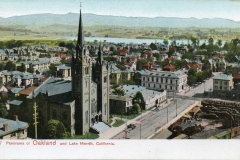 A vintage postcard shows a panoramic view of Oakland, California, featuring a large church with twin spires, surrounding buildings, and Lake Merritt in the background, with mountains in the distance.