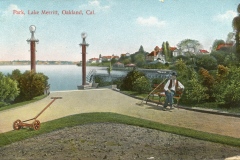 Vintage postcard of Lake Merritt, Oakland, California, featuring a man sitting on a bench with two light poles nearby. A green lawn roller and a hilly, park-like landscape with buildings in the background are visible.