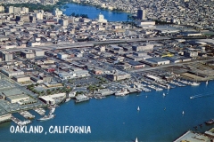 Aerial view of a marina with sailboats in a body of water bordered by industrial and commercial buildings. The cityscape of Oakland, California is visible in the background with a mix of urban structures and greenery. OAKLAND, CALIFORNIA text is overlaid.