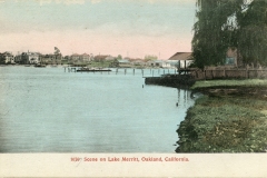 Vintage postcard of Lake Merritt in Oakland, California, featuring calm water, a pier, and a house surrounded by a fence on the right. Trees and more houses line the background, with a pastel sky above.