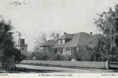 Black and white photo of a historical residence in Livermore, California, featuring a large house with a wraparound porch, surrounded by trees and a picket fence. A windmill is visible in the background.