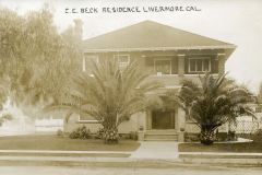 A sepia-toned photo of a two-story house labeled C.E. Beck Residence Livermore Cal. The house is flanked by large palm trees, with a small front yard and a paved walkway leading to the entrance. A chimney is visible on the left.