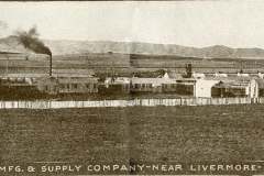 Panoramic view of an early 20th-century industrial facility surrounded by fields and hills. Several buildings with chimneys are visible, emitting smoke. Text reads, The Large Fuse Works of Coast Mfg. & Supply Company near Livermore producing blasting caps and fuses.