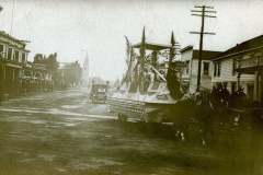 Livermore California, a vintage black and white photo shows a horse-drawn parade float on a street with old wooden buildings and a church steeple in the background. One car is seen ahead, and utility poles line the road. People are on the float.