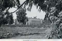 Black and white photo of the Livermore Sanitarium building, partially obscured by trees in the foreground. The structure is surrounded by vegetation, with a lawn and trees in the background.