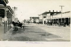 Sepia-toned vintage photo of Main Street, Livermore, California, showing  a dirt road lined with buildings and horse-drawn carriages in a small town. Several horses are hitched to posts along the sidewalk. The scene is bustling with people and shops under a clear sky.
