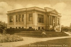 Sepia-toned vintage postcard image of the Livermore Public Library in California. The building features classical architecture with columns and large windows. Two people are seen sitting on the grass in front of the library.