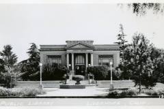 Black and white photograph of a historic public library in Livermore, California. The building features a central arched entrance, columns, and a symmetrical facade. A small fountain is in the foreground, surrounded by trees and streetlamps.