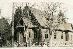 Vintage photograph of a small wooden church with a steep gabled roof and arched windows. Bare trees frame the building, and a picket fence runs in front. The setting suggests early 20th century architecture.
