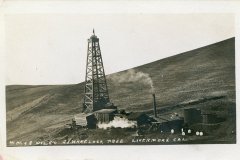 Black and white image of an oil drilling site in a barren landscape. A large wooden oil derrick stands adjacent to small industrial buildings. Smoke rises from a chimney. Text reads WMRS OIL CO J J WHEELOCK PRES LIVERMORE CAL.