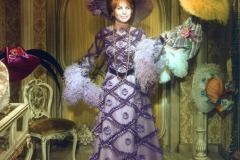 A woman in a lavish purple gown and large matching hat stands in a vintage room decorated with ornate wallpaper and colorful hats. This Barbara Streisand wax figure at Movieland holds a feathery boa and smiles at the camera.