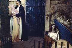 Bella Lugosi as Dracula, leans over a woman in a light dress as if to bite her neck, while a skeleton rises from a coffin nearby in a dimly lit, spooky cemetery scene, at Movieland Wax Museum, Buena Park, California.