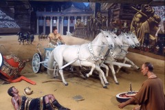 A dramatic scene featuring a Charleton Heston wax figure at Movieland shows a man driving a chariot pulled by four white horses in an arena, with a fallen charioteer and another man kneeling nearby, set against an ancient Roman backdrop.