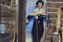 A figure dressed as Charlie Chaplin’s Tramp from The Gold Rush at Movieland Wax Museum stands in a log cabin’s open doorway, cane in hand. Snowshoes, a bucket, and winter gear hang on the rustic wooden walls dusted with snow.