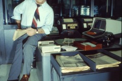 Edward Asner as Lou Grant at Movieland Wax Museum in Buena Park, California, leans on a desk with newspapers, a computer, and Associated Press labels visible, wearing a light shirt and loosened tie, with a serious expression.