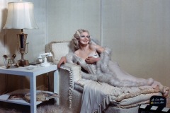 A Jean Harlow wax figure at Movieland lounges in a glamorous white gown and fur stole on a plush chaise, beside a table with a lamp, phone, flowers, and a clapperboard reading Dinner at Eight.